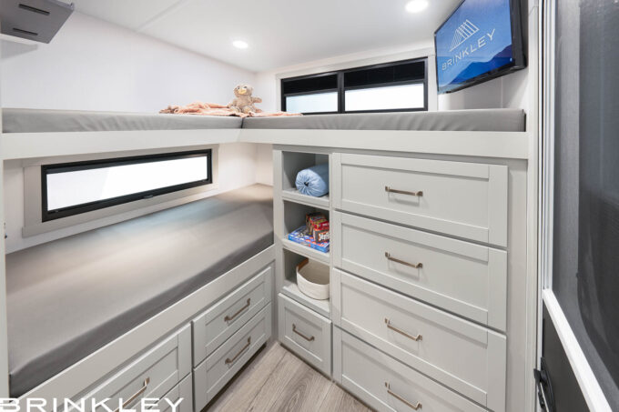 2026 Brinkley Model Z 2810 Fifth Wheel Rear Bunkhouse Interior featuring triple bunk beds, extensive white cabinetry with deep storage drawers and cubbies, a wall-mounted smart TV, and a private off-campside entry door.