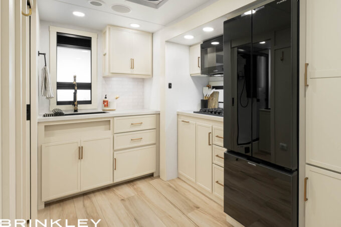 Kitchen interior of the Brinkley Model I 235 Travel Trailer showcasing high-quality construction details found in the best built travel trailers. Features include top-grade hardwood cabinetry with durable dovetail drawers, solid surface countertops, and an oversized stainless steel farmhouse sink. The space is illuminated by industry-exclusive square frameless Euro-styled windows with CraftSense™ hardwood trim, offering a residential look and superior ventilation.